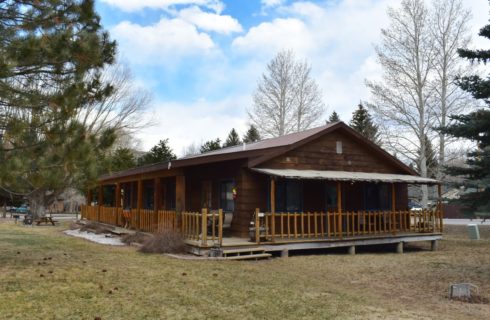 Large wooden lodge with blue painted trim surrounded by large pine trees and green grass