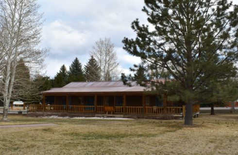 Large wooden lodge with blue painted trim and wrap around porch surrounded by large pine trees and green grass