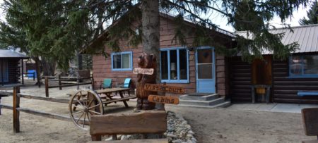 Wooden cottage with blue painted door and trim surrounded by large evergreen trees with a picnic table out front