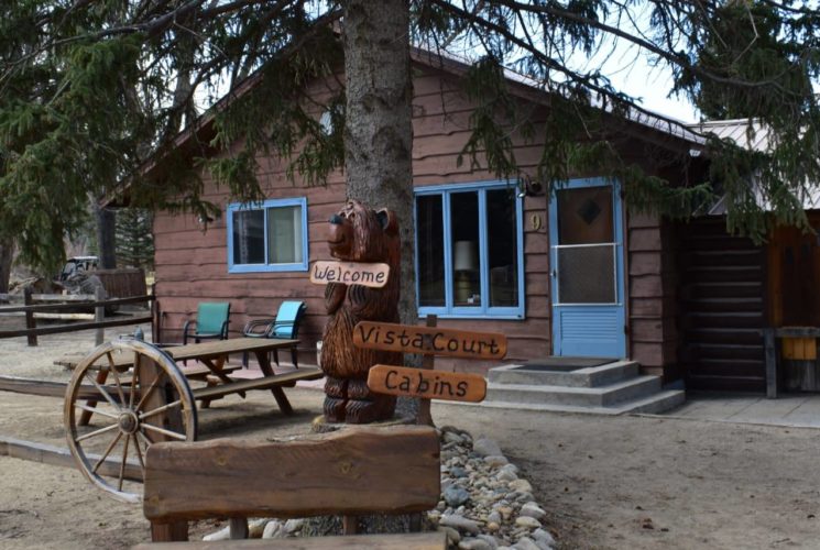 Wooden cottage with blue painted door and trim surrounded by large evergreen trees with a picnic table out front