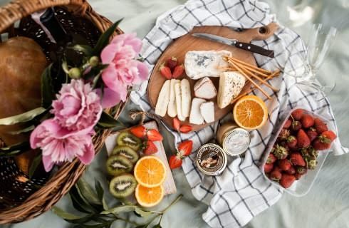 Top view of picnic basket with slicked kiwi, oranges, strawberries and cheese on towel nearby